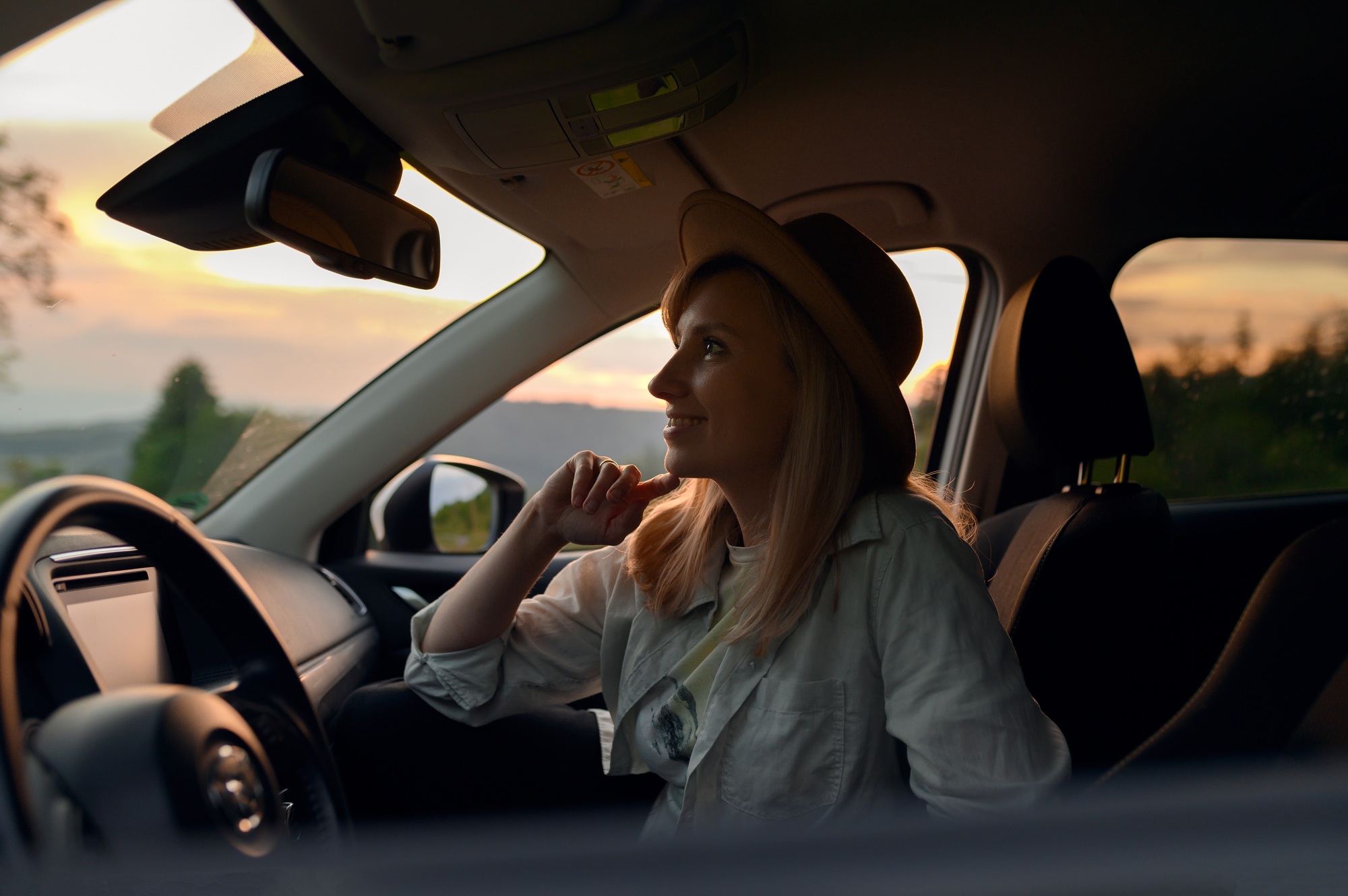 Smiling and happy young blonde woman in hat sitting in a rental car on vacations